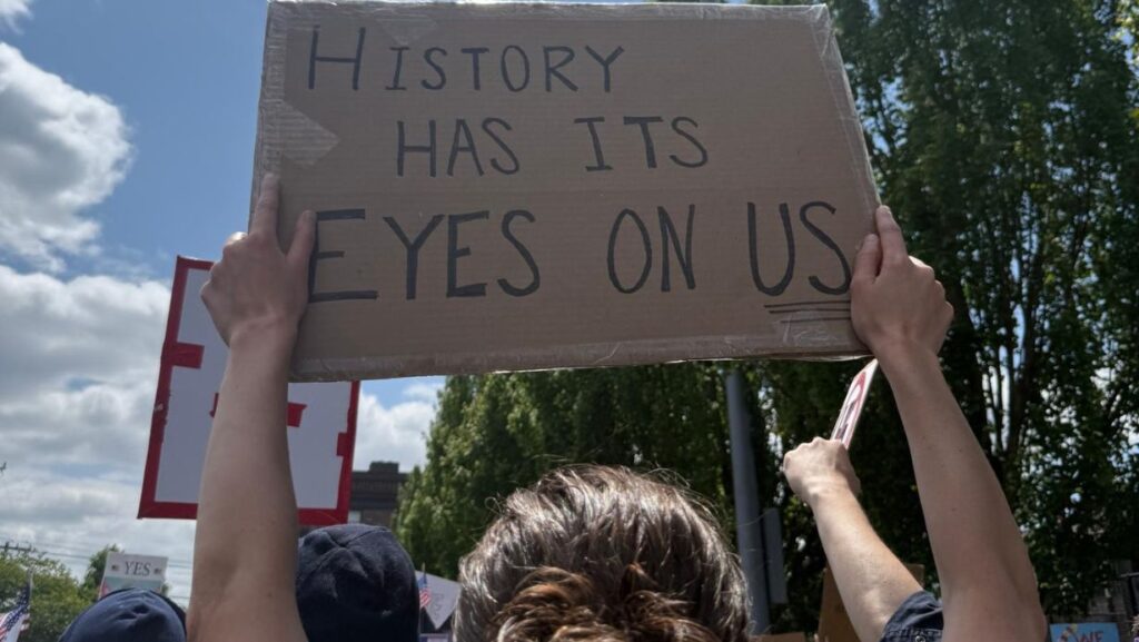 Una manifestante sostiene un cartel con la frase “History has its eyes on us” durante la protesta, subrayando la importancia histórica del momento. Foto: para Latino Herald Maia Vázquez