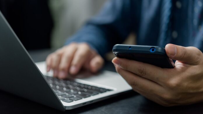 Hombre sostiene un celular y escribe en su computadora