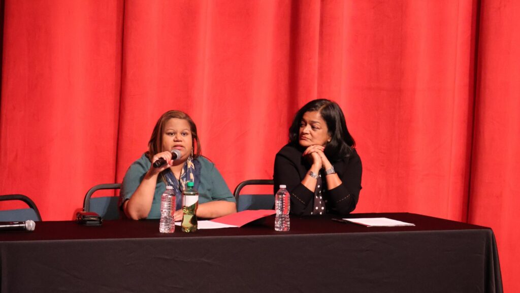 Shasti Conrad hablando al micrófono mientras Pramila Jayapal la observa sentada en una mesa durante el Town Hall. © Latino Herald