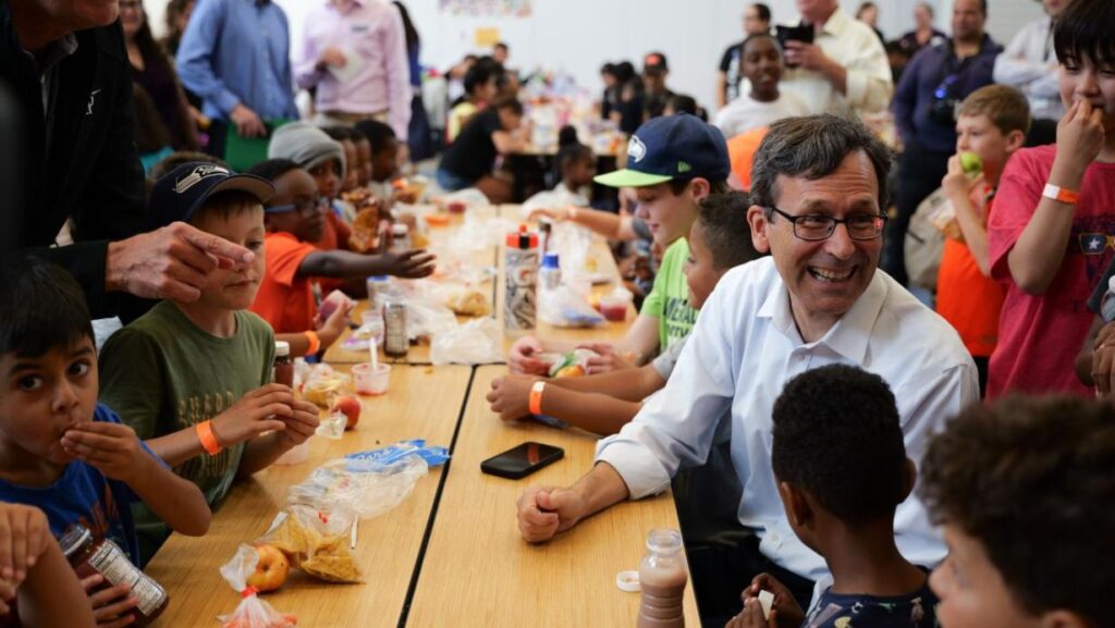 l gobernador Bob Ferguson sonríe mientras conversa con niños en el comedor de la escuela Lynnwood Elementary, en el marco de un programa de comidas de verano relacionado con discusiones sobre SNAP. (Foto cortesía de la oficina del gobernador de Washington, 08/21/2025)