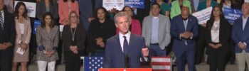 El gobernador de California, Gavin Newsom, ofrece un discurso sobre la redistribución de mapas electorales frente a un grupo de legisladores y líderes demócratas sosteniendo carteles de “Defend Democracy” y “Election Rigging Response Act”.