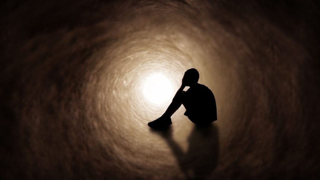 Silueta de una persona sentada sola en un túnel oscuro, con una pequeña luz al final. Representa los sentimientos de desesperación y soledad.