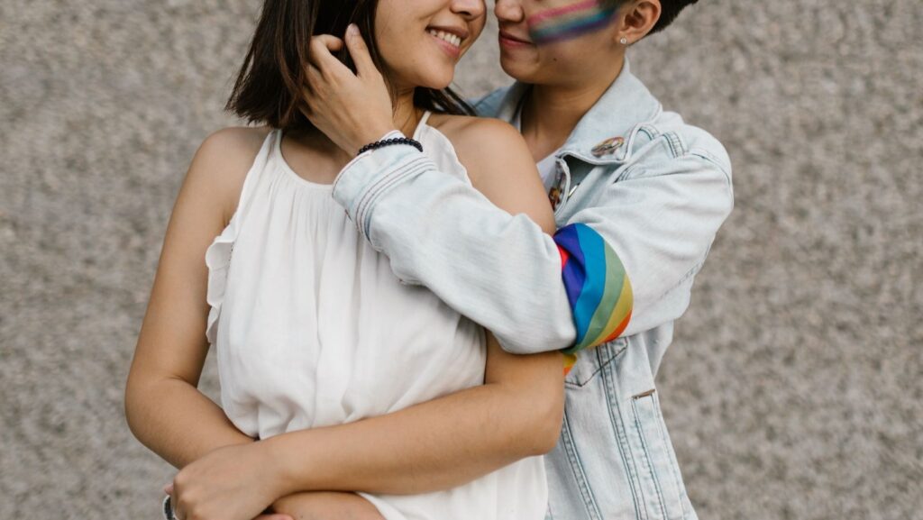Dos personas sonriendo y abrazándose afectuosamente, una de ellas lleva maquillaje con los colores de la bandera del arcoíris en la mejilla y una banda con los mismos colores en la manga.