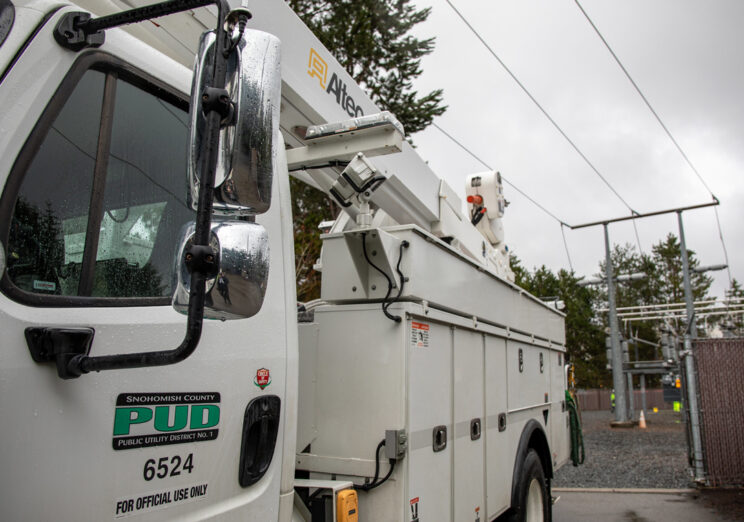 Parte delantera de un camión de servicio de Snohomish County PUD bajo una lluvia ligera. El logotipo de la PUD es visible en la puerta, con un brazo de la grúa extendido hacia el fondo de la imagen, donde hay equipos de la red eléctrica.