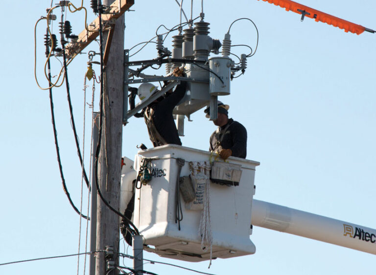  Dos trabajadores de servicios públicos en una grúa, instalando un recloser inteligente en un poste de la red eléctrica. Llevan cascos de seguridad y están colocando el nuevo equipo para modernizar el sistema.
