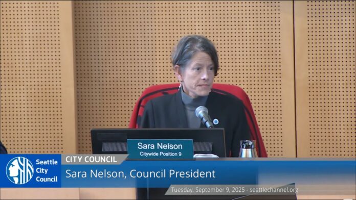 1 Sara Nelson, presidenta del Concejo de Seattle, sentada frente a un micrófono durante la reunión del concejo, con un letrero que indica su nombre y cargo.