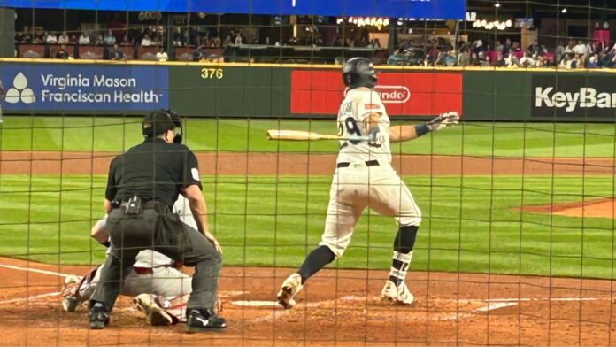 22 GENERAL (4) Un jugador de béisbol con un uniforme crema y rayas azules conecta un hit. Se ve desde atrás al catcher en cuclillas y al umpire gritando la jugada.