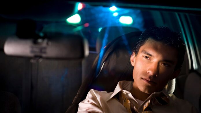 Hombre hispano en el asiento de un vehículo con luces rojas y azules de un vehículo policial detrás de él.