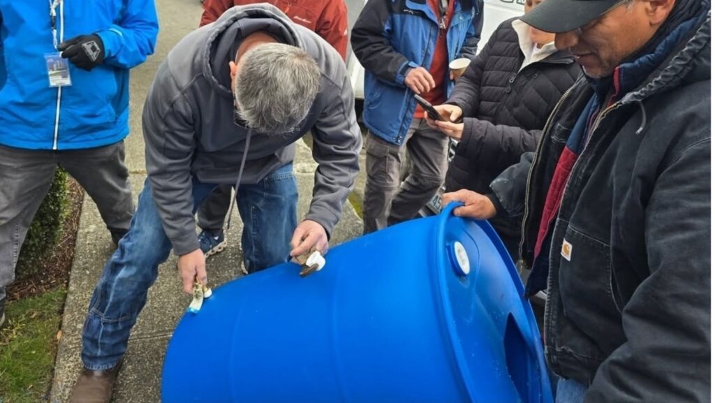 Un grupo de personas observa a un hombre arrodillado que instala una válvula en la base de un gran barril de plástico azul. Están al aire libre, en un espacio de estacionamiento.