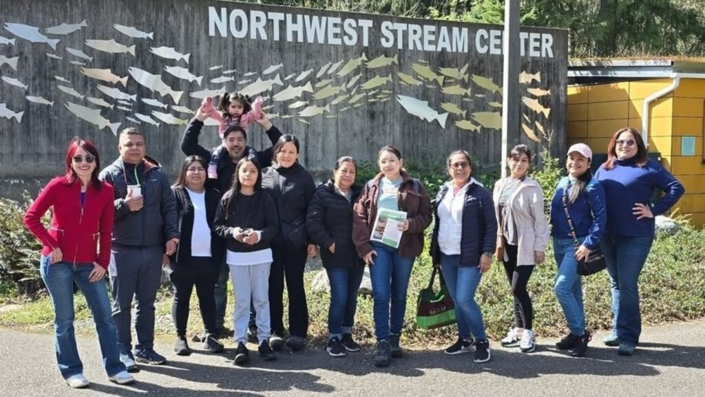 Un grupo de personas sonríe y posa para una foto frente a la entrada del "Northwest Stream Center", con un mural de peces en la pared detrás de ellos.