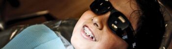 Primer plano de un niño con gafas de sol protectoras recostado en un sillón dental, sonriendo levemente, mostrando frenos.