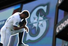 Octubre es proclamado como el Mes de los Mariners en Seattle Jugador de los Seattle Mariners arrodillado en el campo con la gorra en el rostro, frente a una gran pantalla que muestra el logotipo del equipo.