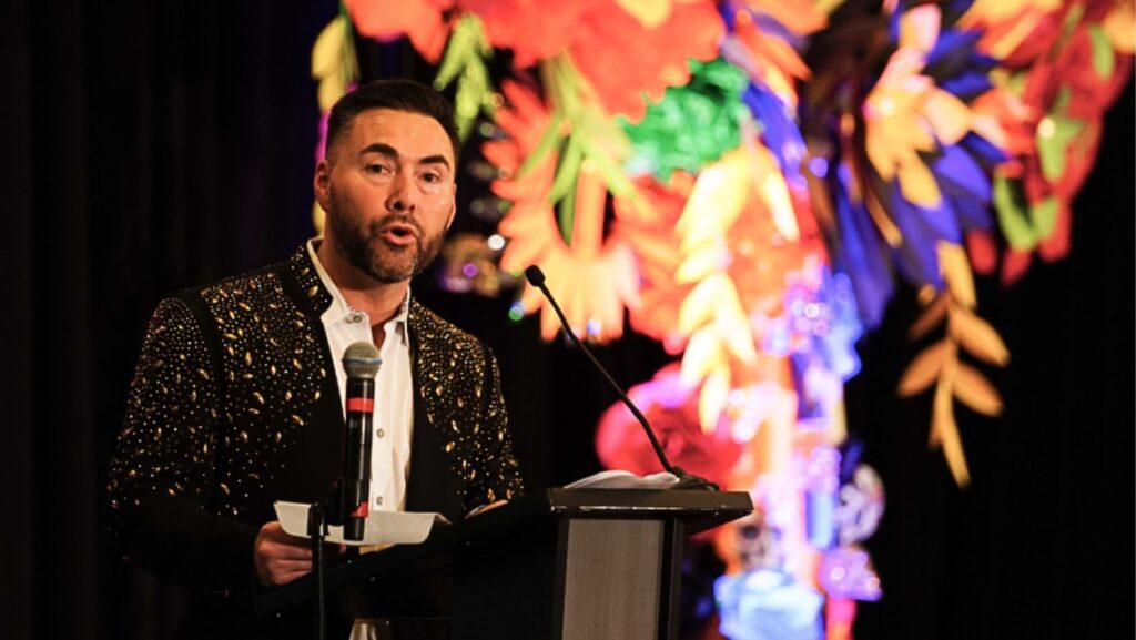Un presentador habla en la gala anual de Entre Hermanos frente a una colorida decoración.