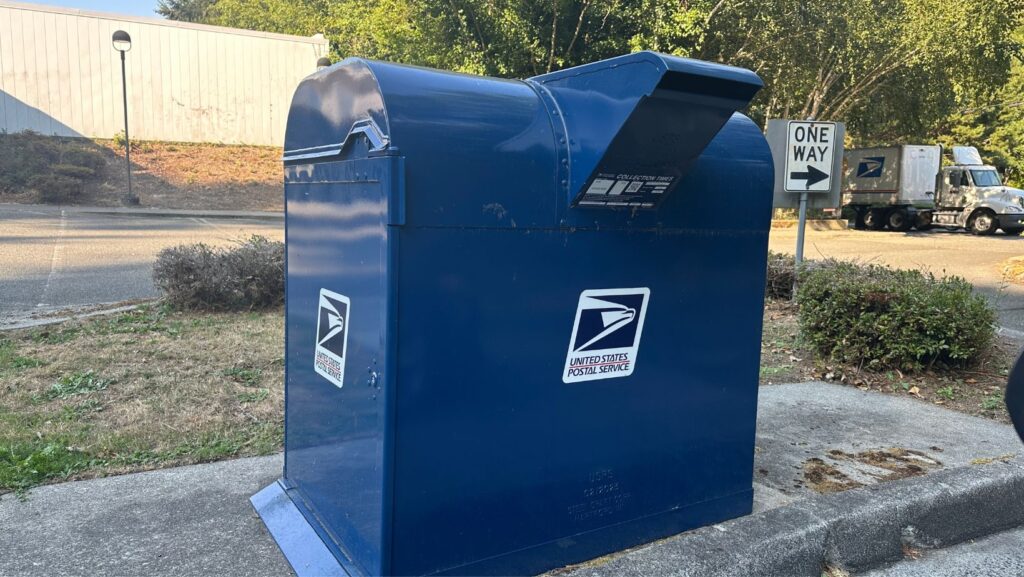 Buzón azul del Servicio Postal de Estados Unidos (USPS) ubicado en una acera, con letrero de “One Way” y un camión postal al fondo en un entorno urbano con árboles.