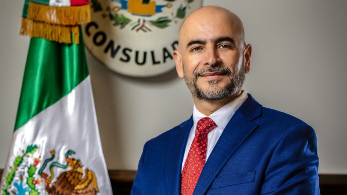 Víctor Hugo López, Cónsul de México en Seattle, de pie en la sede consular con la bandera de México al fondo.