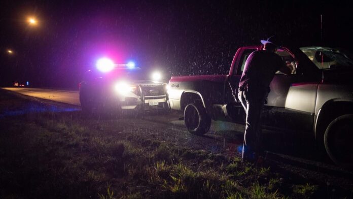 Oficial de la Patrulla Estatal de WA detiene a un conductor en una carretera oscura bajo la lluvia, con luces policiales encendidas