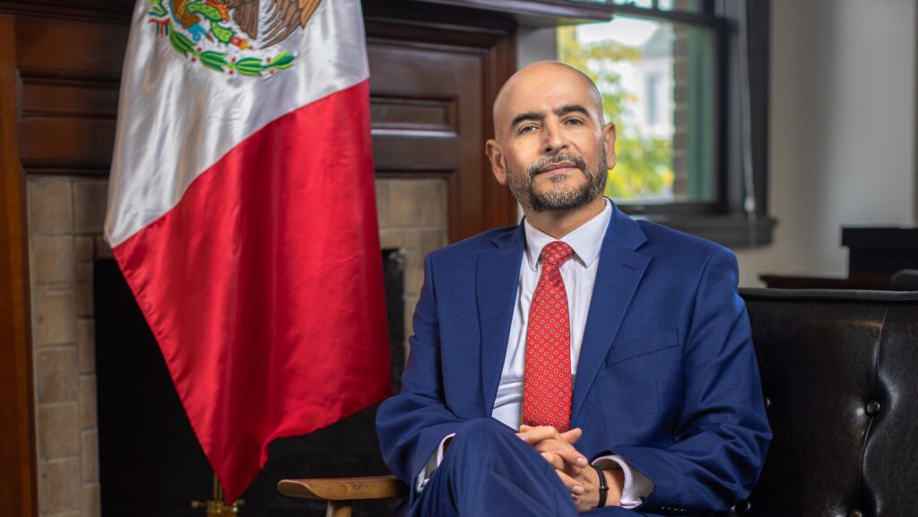 Víctor Hugo López, Cónsul de México en Seattle, sentado en oficina del consulado junto a la bandera de México.