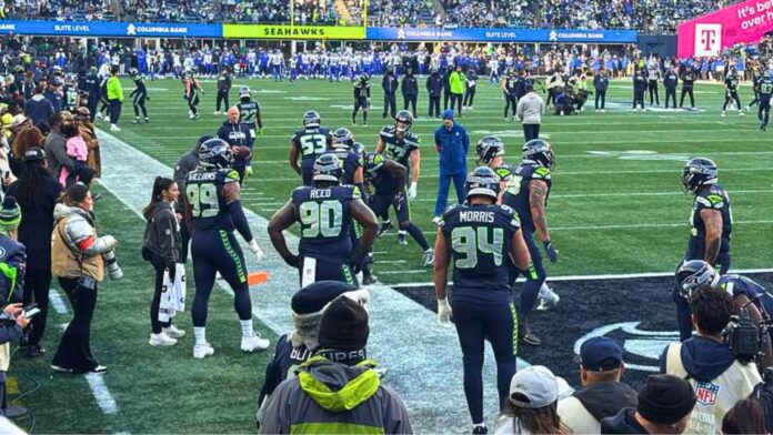 Seattle Seahawks realizando ejercicios de calentamiento en la línea lateral del campo en el estadio Lumen Field antes del partido contra Los Angeles Rams, rodeados de aficionados y personal del equipo.