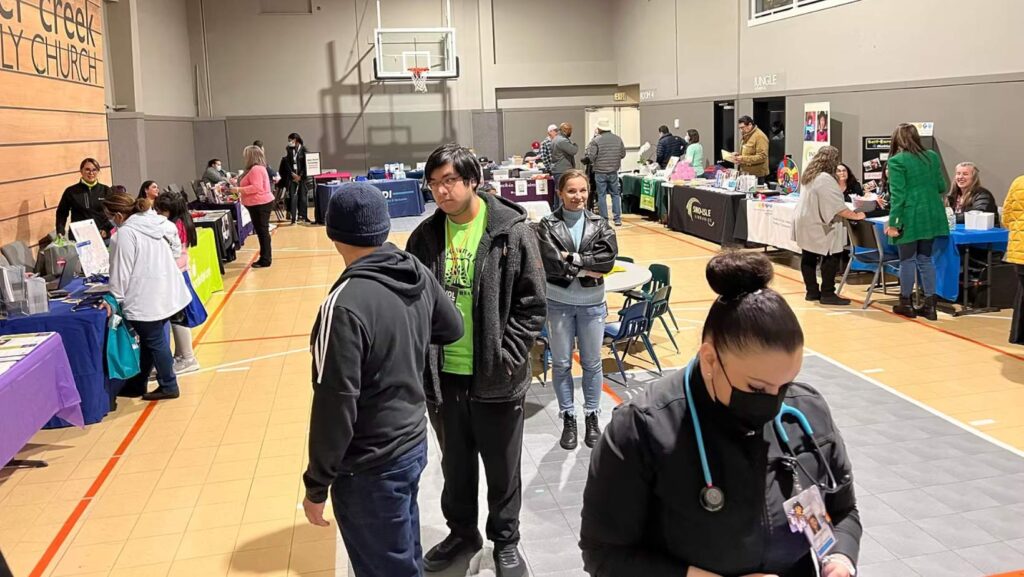 Personas recorren una feria comunitaria de servicios de salud instalada en un gimnasio, con mesas informativas de distintas organizaciones y personal médico atendiendo al público.
