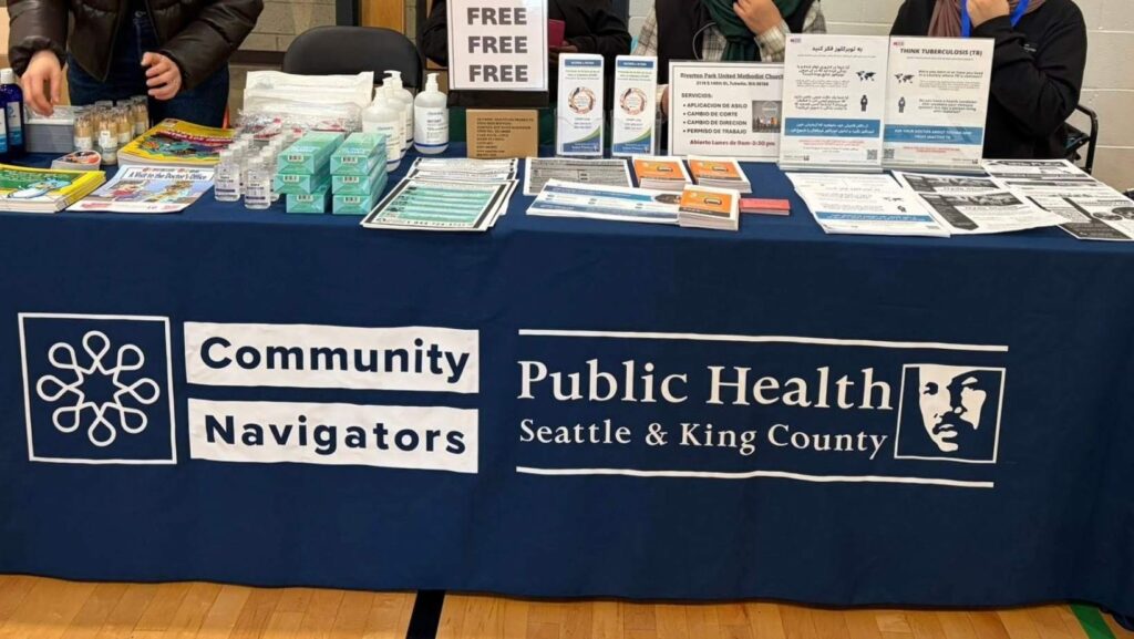 Mesa informativa de Community Navigators y Public Health Seattle & King County con folletos bilingües, pruebas de salud gratuitas y materiales educativos sobre servicios comunitarios y prevención de enfermedades.
