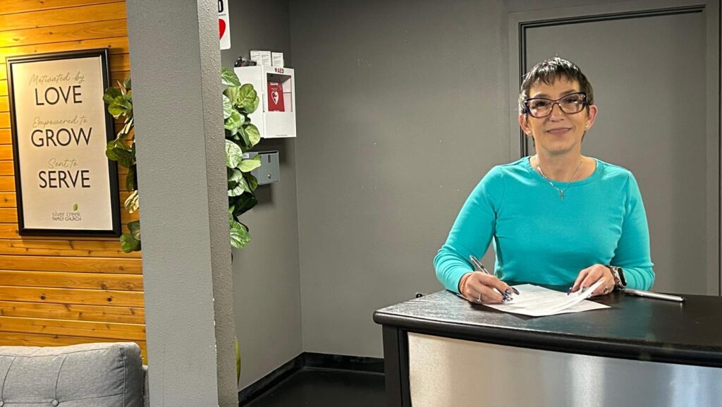 Mujer de pie en un mostrador revisa y firma documentos en el interior de un edificio comunitario, con un mensaje motivacional visible en la pared