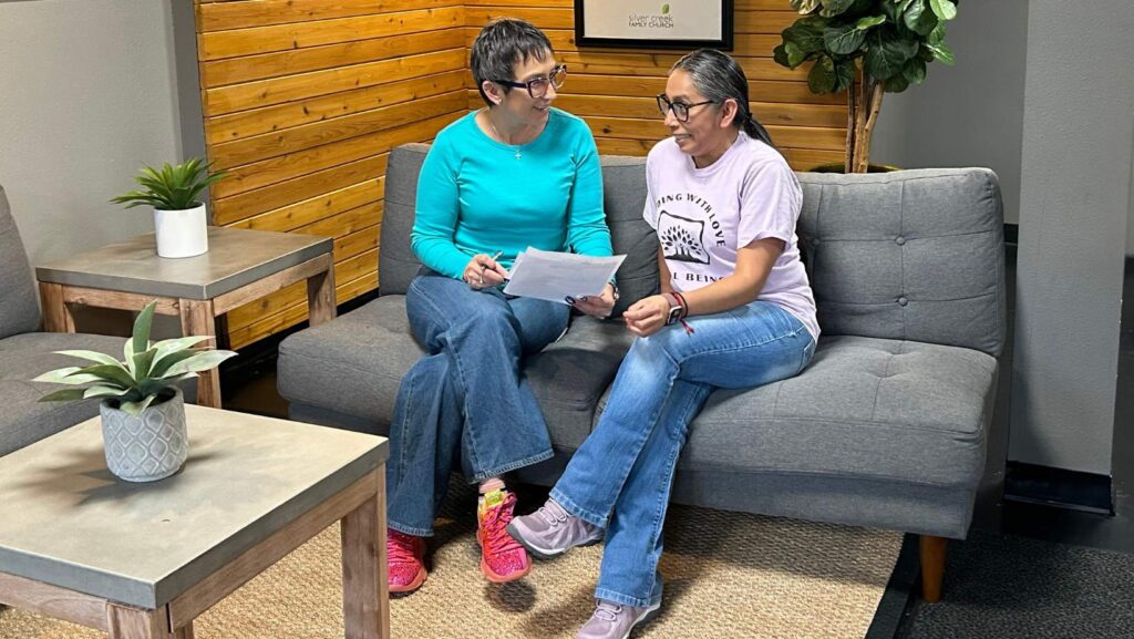 Dos mujeres sentadas en un sofá revisan documentos juntas dentro de un espacio comunitario, en un ambiente de acompañamiento y apoyo.