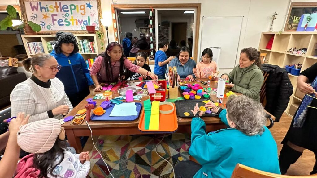 Adultos y niños participan en una actividad manual alrededor de una mesa, creando figuras coloridas con materiales de arte en un espacio comunitario.