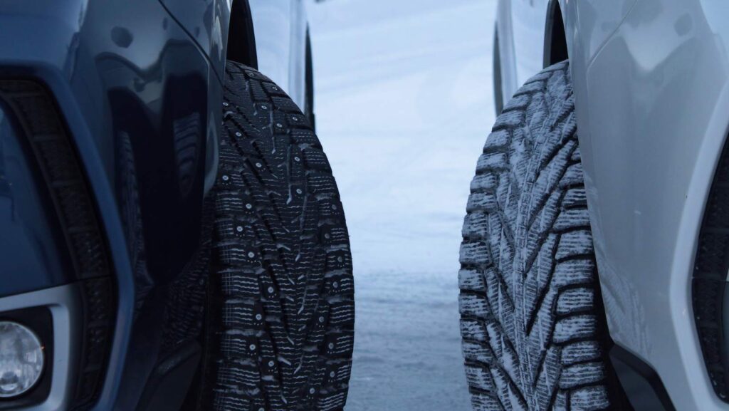 Primer plano de llantas de vehículos sobre una superficie nevada: a la izquierda, una llanta con tacos metálicos visibles; a la derecha, una llanta de invierno con dibujo profundo cubierta de nieve.