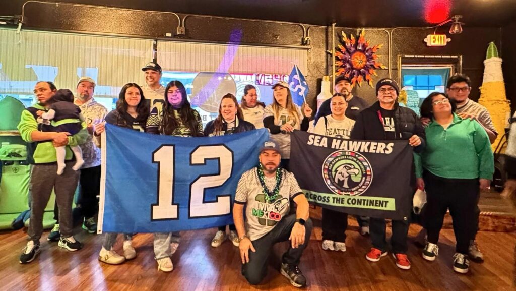 Alt text: Grupo de aficionados de Sea Hawkers Latin America reunidos en un restaurante sosteniendo una bandera con el número 12 de los Seahawks