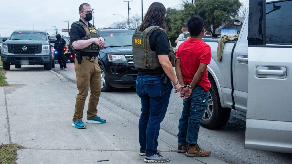Agentes de HSI detienen a un hombre esposado junto a una camioneta blanca durante un operativo en la calle.