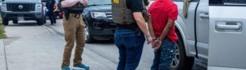 Agentes de HSI detienen a un hombre esposado junto a una camioneta blanca durante un operativo en la calle.