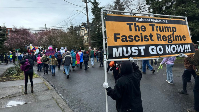 Manifestantes marchan por una calle de Seattle durante la Marcha de Mujeres del 8 de marzo mientras una persona sostiene un gran cartel de protesta contra Donald Trump entre la multitud.