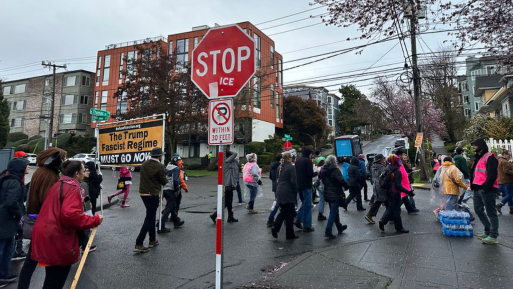 Manifestantes cruzan una intersección en Seattle durante la Marcha de Mujeres del 8 de marzo, con pancartas y un letrero que dice “Stop ICE”, mientras avanzan hacia el Parque Cal Anderson.