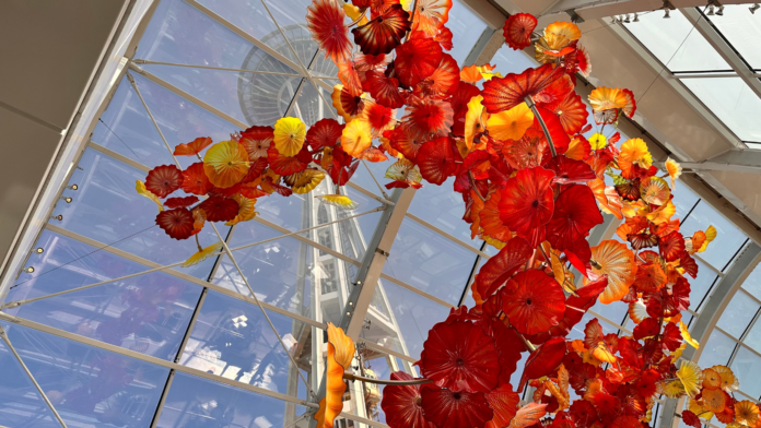 Arte en vidrio soplado con formas florales en tonos rojos, naranjas y amarillos suspendida en el techo de un invernadero con estructura de vidrio en Chihuly Garden and Glass.