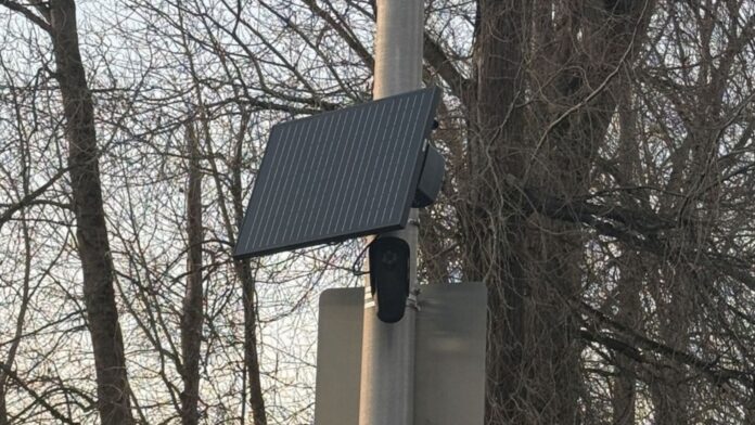 Cámara de seguridad Flock con panel solar instalada en un poste en Renton, rodeada de árboles.