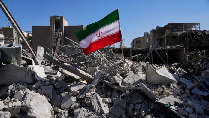 Bandera iraní ondea entre escombros Una bandera iraní ondea entre los escombros de una estación de policía destruida tras los bombardeos de Estados Unidos e Israel en Teherán, Irán, en marzo de 2026 (Foto de AP/Vahid Salemi)