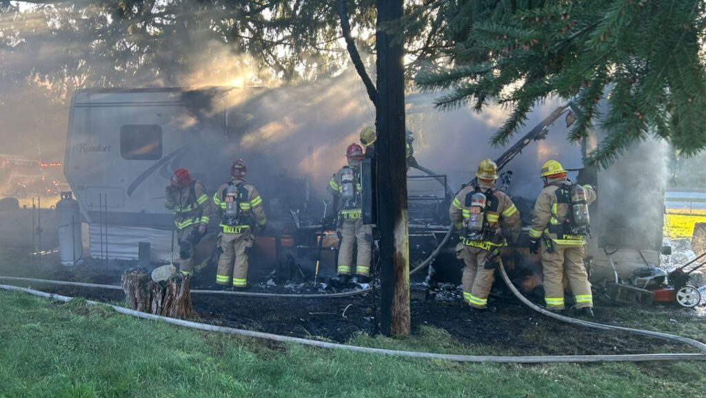 Bomberos trabajan alrededor de un tráiler humeante en Woodinville, mientras controlan los restos del incendio entre árboles y equipo de emergencia.