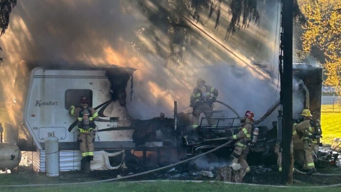 Bomberos combaten un incendio activo en un tráiler en Woodinville, rodeado de humo espeso y llamas visibles, mientras trabajan cerca de un tanque de propano y postes eléctricos.