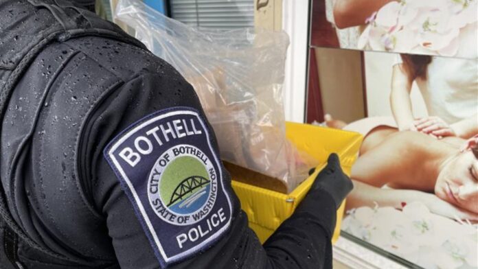 Agente de la policía de Bothell sostiene una caja con evidencia frente a un establecimiento de masajes durante un operativo.