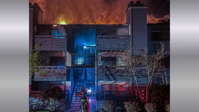 Bombero frente a un edificio de apartamentos de dos pisos envuelto en llamas durante la noche, con fuego intenso saliendo del techo, humo denso en el cielo y luces de emergencia iluminando la escena.
