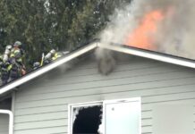 Incendio en Lynnwood daña townhomes y deja familias sin hogar Bomberos sobre el techo de una vivienda combaten un incendio activo mientras llamas y humo denso salen del ático; una ventana inferior está rota y la estructura presenta daños visibles.