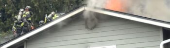 Bomberos sobre el techo de una vivienda combaten un incendio activo mientras llamas y humo denso salen del ático; una ventana inferior está rota y la estructura presenta daños visibles.