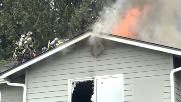 Bomberos sobre el techo de una vivienda combaten un incendio activo mientras llamas y humo denso salen del ático; una ventana inferior está rota y la estructura presenta daños visibles.