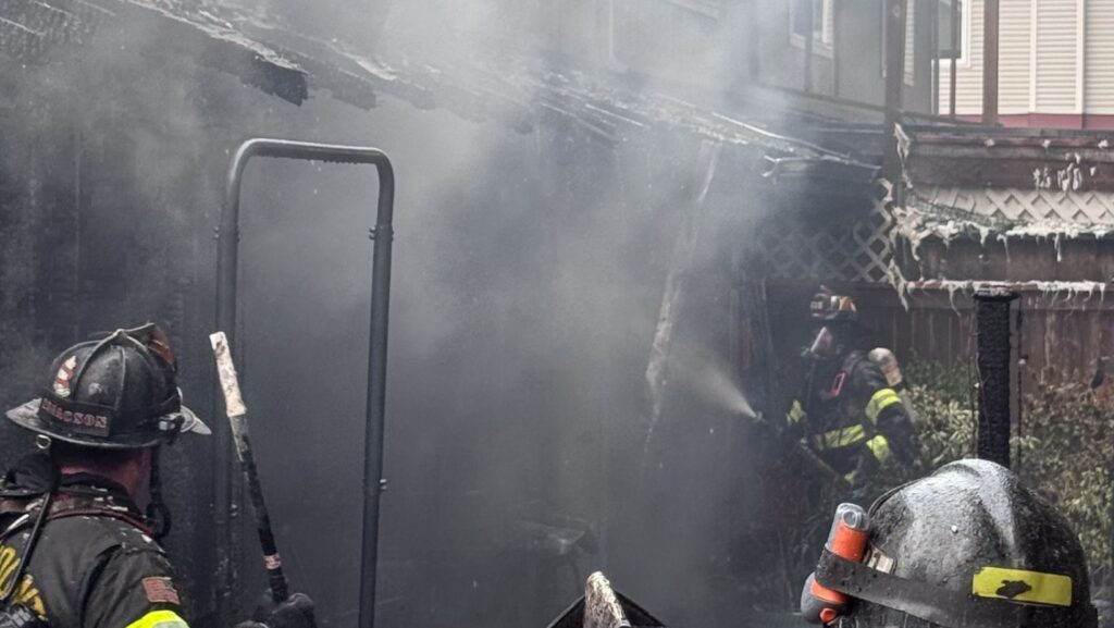 Bomberos combaten un incendio en la parte trasera de una vivienda mientras una densa columna de humo cubre la estructura; el exterior aparece severamente dañado y ennegrecido por las llamas.