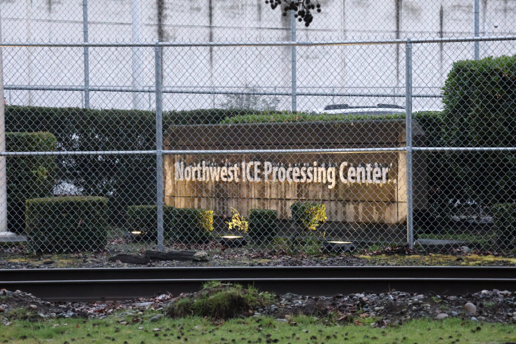 Vista exterior del Northwest ICE Processing Center en Tacoma, Washington, detrás de una cerca de seguridad con alambre; se observa el letrero de la instalación y vías de tren en primer plano.