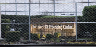 Vista exterior del Northwest ICE Processing Center en Tacoma, Washington, detrás de una cerca de seguridad con alambre; se observa el letrero de la instalación y vías de tren en primer plano.