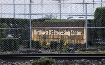 Juez autoriza inspecciones sanitarias en el centro de detención de GEO en Tacoma Vista exterior del Northwest ICE Processing Center en Tacoma, Washington, detrás de una cerca de seguridad con alambre; se observa el letrero de la instalación y vías de tren en primer plano.