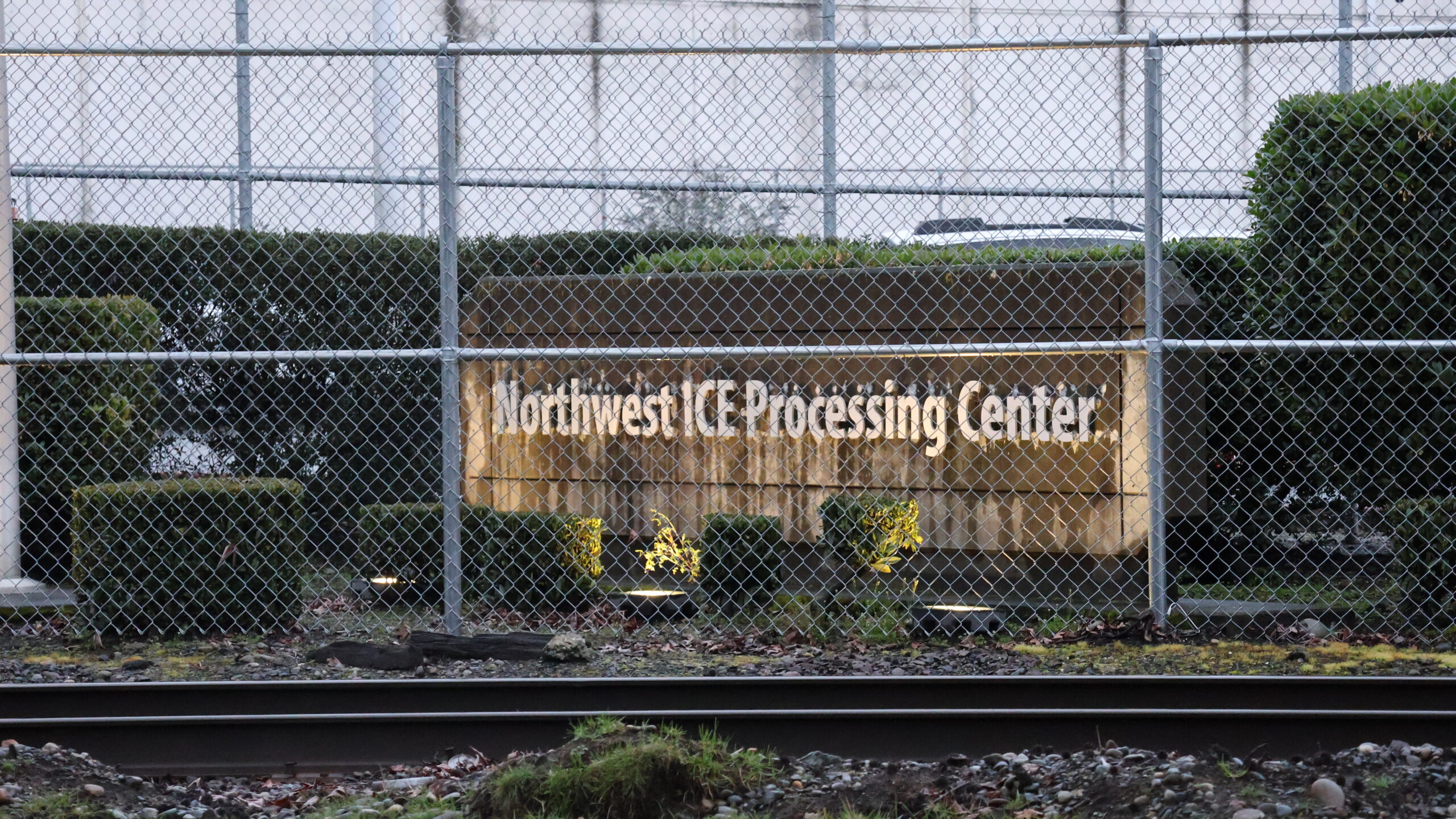 Vista exterior del Northwest ICE Processing Center en Tacoma, Washington, detrás de una cerca de seguridad con alambre; se observa el letrero de la instalación y vías de tren en primer plano.