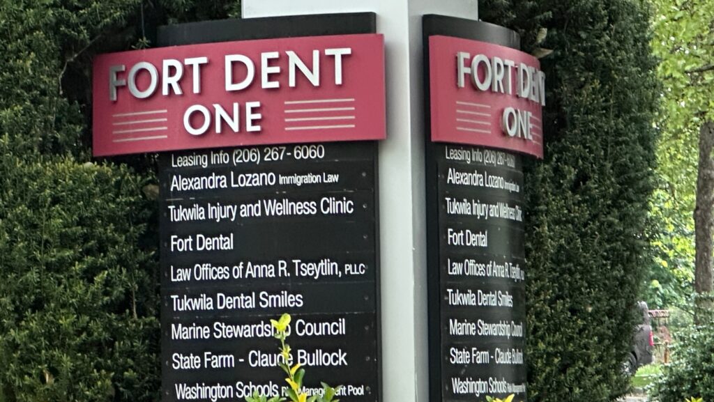 Letrero de directorio comercial en el complejo Fort Dent One, en Tukwila, Washington, donde aparece el nombre de Alexandra Lozano Immigration Law entre los negocios que operan en el edificio. La imagen muestra jardines ornamentales y áreas verdes que rodean la entrada del complejo.