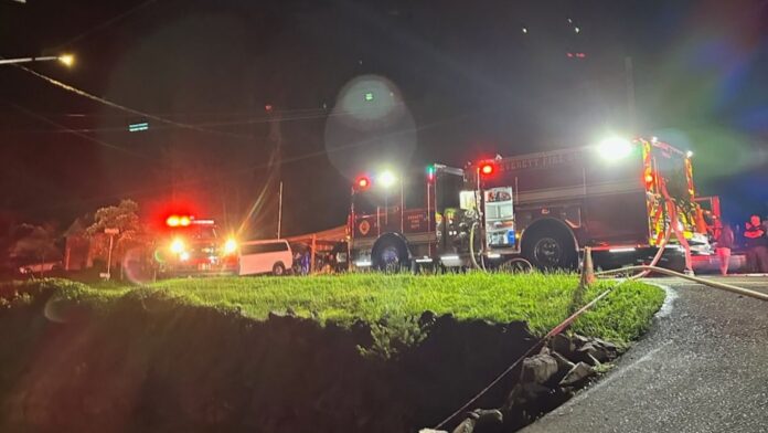 Equipos de emergencia Camión de bomberos y unidades de emergencia con luces rojas encendidas durante la noche, mientras mangueras están desplegadas en una calle residencial iluminada.